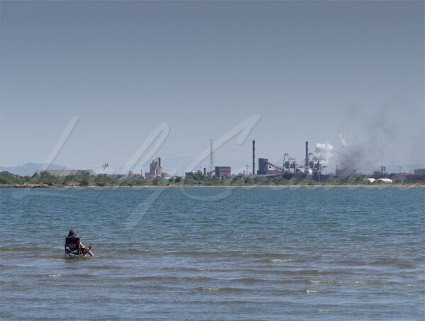 A bather in search of freshness in her partially submerged seat facing industrial installations.